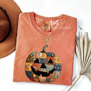 May include: A coral-coloured long-sleeve shirt featuring a patchwork pumpkin design. The pumpkin has a black face and is made of various floral and patterned fabrics. The shirt has a crew neck and a tag that reads "MAKES CUSTOMIZED".