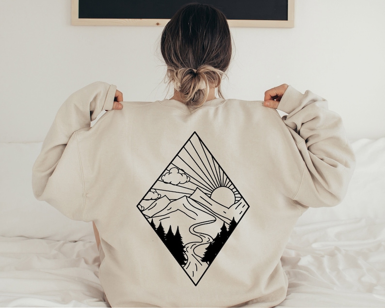 Puede incluir: Una sudadera de color crema con un dibujo de l&iacute;nea negra de un paisaje de monta&ntilde;a con un sol y nubes. El dise&ntilde;o tiene forma de diamante.