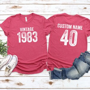 K&ouml;nnte beinhalten: Zwei rote T-Shirts mit wei&szlig;em Text. Das vordere Shirt sagt "Vintage 1983". Das hintere Shirt sagt "Custom Name 40".