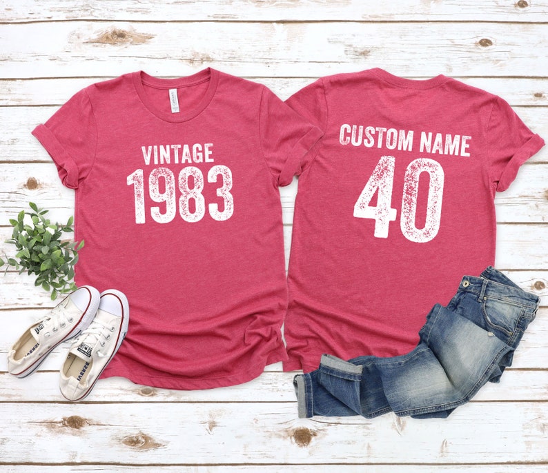 K&ouml;nnte beinhalten: Zwei rote T-Shirts mit wei&szlig;em Text. Das vordere Shirt sagt "Vintage 1983". Das hintere Shirt sagt "Custom Name 40".