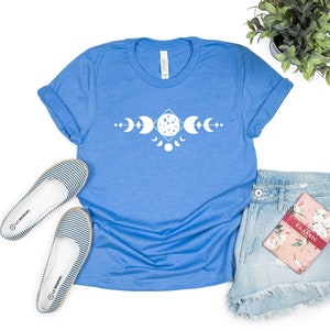 May include: A blue t-shirt with a white moon phases design. The shirt is laid flat on a white background with a pair of striped flats, denim shorts, and a floral notebook.