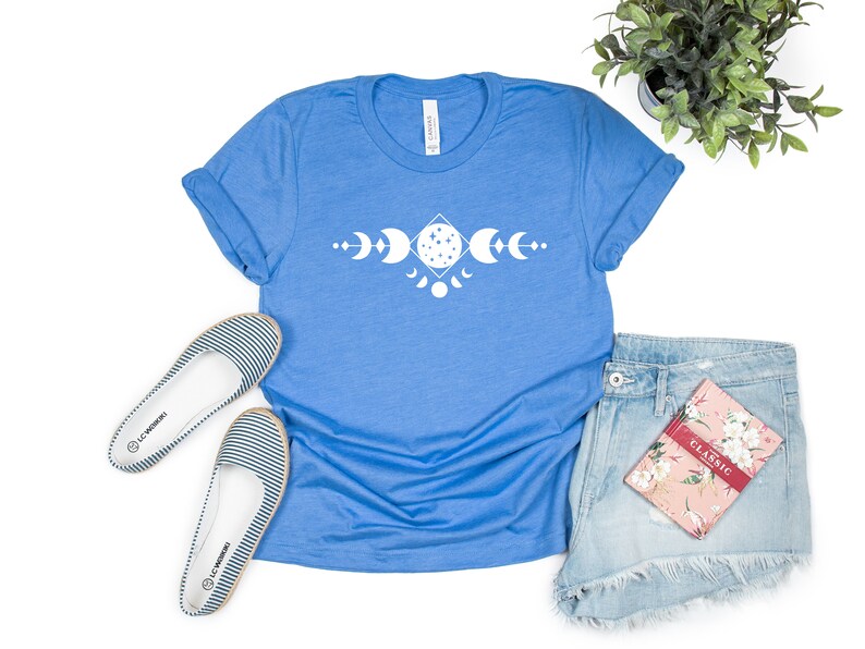 May include: A blue t-shirt with a white moon phases design. The shirt is laid flat on a white background with a pair of striped flats, denim shorts, and a floral notebook.