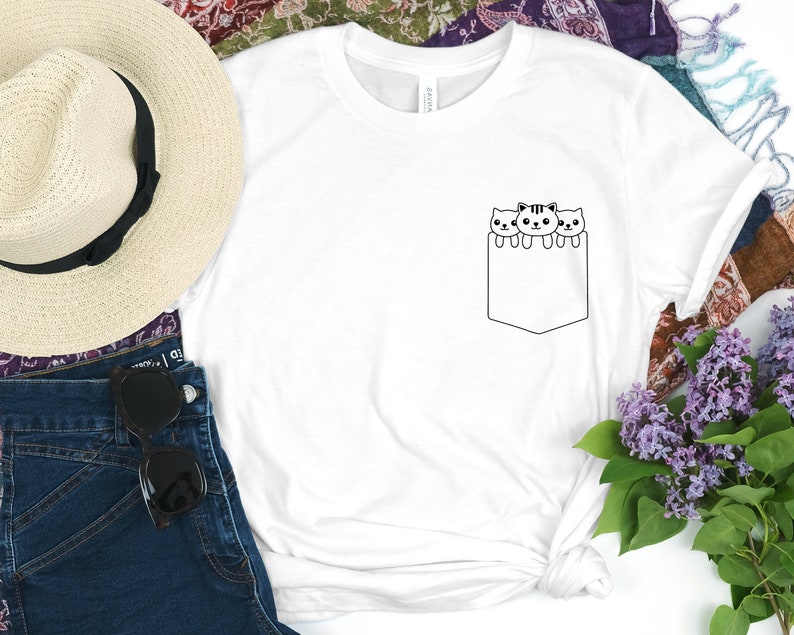 May include: White t-shirt with a black outline of three cats peeking out of a pocket on the front of the shirt.