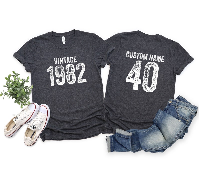 May include: Two dark gray t-shirts with white text. One shirt says "Vintage 1982" and the other says "Custom Name 40".