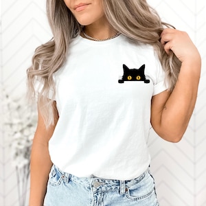 May include: White t-shirt with a black cat peeking out from behind the fabric. The cat has yellow eyes.