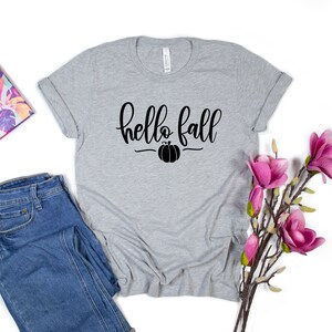 May include: A gray t-shirt with the text "hello fall" in black script with a small pumpkin graphic underneath. The shirt is laid flat on a white background with a pair of blue jeans and a floral planner to the left and a sprig of pink flowers to the right.