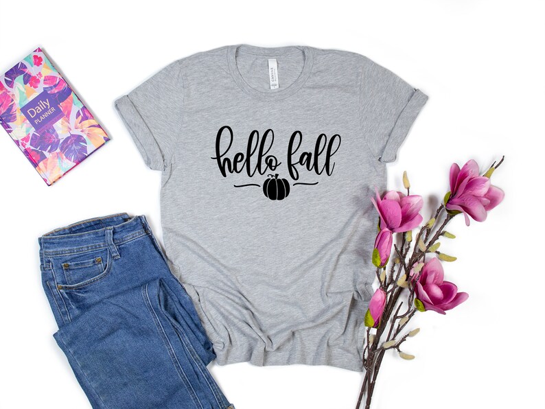 May include: A gray t-shirt with the text "hello fall" in black script with a small pumpkin graphic underneath. The shirt is laid flat on a white background with a pair of blue jeans and a floral planner to the left and a sprig of pink flowers to the right.