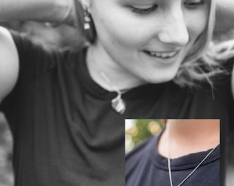 Peut inclure: Un collier composé d'un pendentif en pierre lisse de couleur ambre en forme de goutte, entouré d'un fin fil métallique argenté. Le pendentif est suspendu à une chaîne fine et simple reposant sur un t-shirt sombre, mettant en valeur le design.