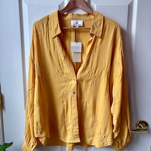 May include: Mustard yellow button-down shirt with long sleeves, hanging on a wooden hanger. The shirt features embroidered floral patterns and a tag. The shirt is made of a soft, flowing fabric.