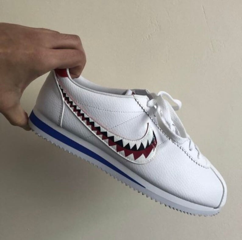 Pu&ograve; includere: Sneakers bianche in pelle con bordino blu e un design a dente di squalo rosso, nero e bianco sul lato.