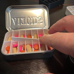 Puede incluir: Una caja de p&iacute;ldoras de plata con la palabra "VITOLD2" grabada en la tapa. La caja est&aacute; abierta y llena de p&iacute;ldoras de varios colores, incluyendo naranja, rosa y blanco.