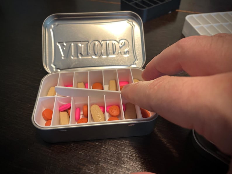 Puede incluir: Una caja de p&iacute;ldoras de plata con la palabra "VITOLD2" grabada en la tapa. La caja est&aacute; abierta y llena de p&iacute;ldoras de varios colores, incluyendo naranja, rosa y blanco.