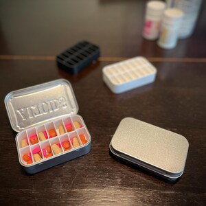 Puede incluir: Una caja de p&iacute;ldoras de metal plateado con la palabra "YMOD2" impresa en la tapa. La caja est&aacute; abierta y llena de p&iacute;ldoras rosas y naranjas. Hay una segunda caja de p&iacute;ldoras de metal plateado vac&iacute;a al lado.