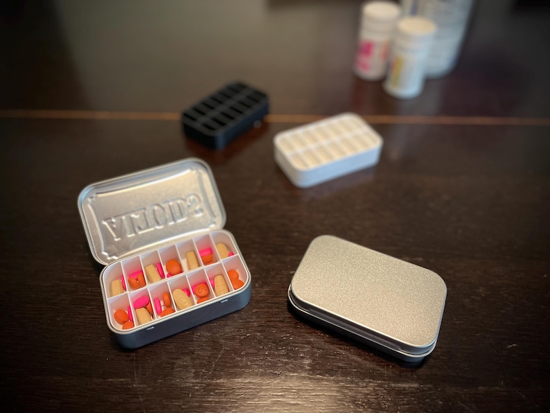 Puede incluir: Una caja de p&iacute;ldoras de metal plateado con la palabra "YMOD2" impresa en la tapa. La caja est&aacute; abierta y llena de p&iacute;ldoras rosas y naranjas. Hay una segunda caja de p&iacute;ldoras de metal plateado vac&iacute;a al lado.
