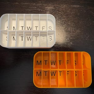 Puede incluir: Dos organizadores de pastillas, uno blanco y otro naranja, con compartimentos etiquetados para cada d&iacute;a de la semana. El organizador blanco tiene los d&iacute;as de la semana escritos, mientras que el organizador naranja usa abreviaturas.