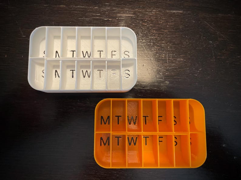 Puede incluir: Dos organizadores de pastillas, uno blanco y otro naranja, con compartimentos etiquetados para cada d&iacute;a de la semana. El organizador blanco tiene los d&iacute;as de la semana escritos, mientras que el organizador naranja usa abreviaturas.
