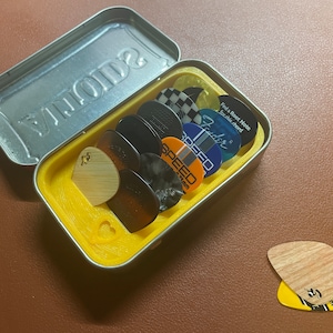 May include: A collection of guitar picks in a silver tin. The picks are various colors and designs, including wood grain, checkered, and branded with "Fender" and "SPEED". The tin has a yellow interior and the word "SONOMA" embossed on the lid.