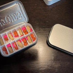 Puede incluir: Una caja de p&iacute;ldoras de metal plateado con la palabra "VIIOIDS?" impresa en la tapa. La caja est&aacute; abierta y contiene una variedad de p&iacute;ldoras de color rosa y naranja.