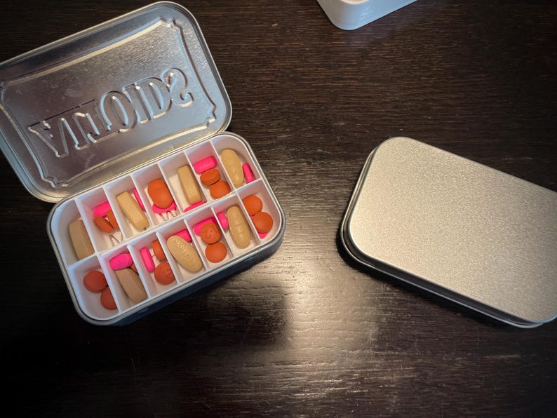 Puede incluir: Una caja de p&iacute;ldoras de metal plateado con la palabra "VIIOIDS?" impresa en la tapa. La caja est&aacute; abierta y contiene una variedad de p&iacute;ldoras de color rosa y naranja.