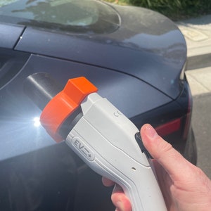 May include: A white electric vehicle charging handle with an orange safety latch. The handle has a black button and the text "EV=+" is printed on the side.