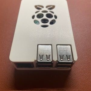 May include: White plastic case for a Raspberry Pi computer. The case has a cut-out for the Raspberry Pi logo and three USB ports.