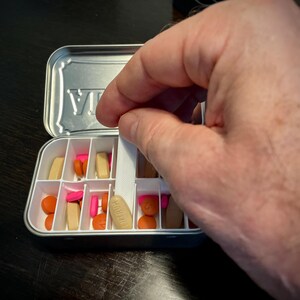 Puede incluir: Un organizador de pastillas blanco con una tapa plateada. La tapa tiene la palabra "VIA" impresa. El organizador est&aacute; lleno de pastillas de varios colores, incluyendo rosa, naranja y marr&oacute;n.