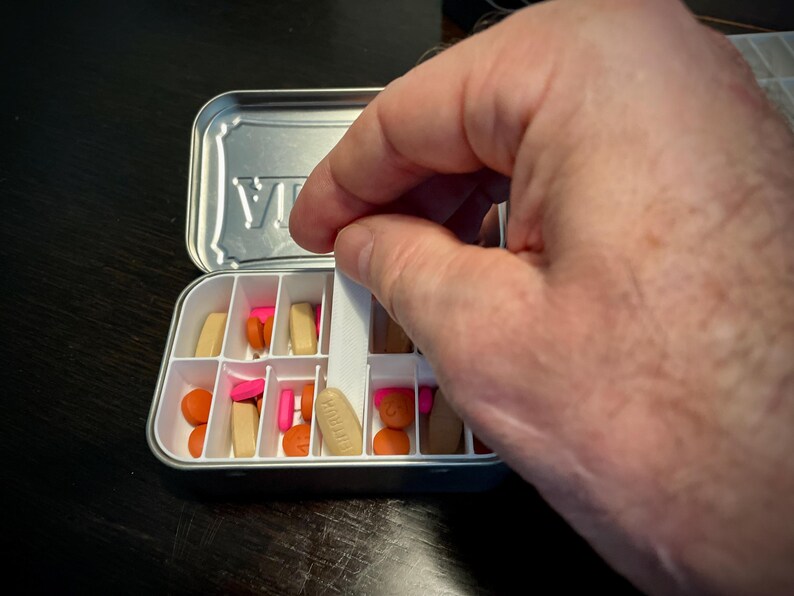 Puede incluir: Un organizador de pastillas blanco con una tapa plateada. La tapa tiene la palabra "VIA" impresa. El organizador est&aacute; lleno de pastillas de varios colores, incluyendo rosa, naranja y marr&oacute;n.