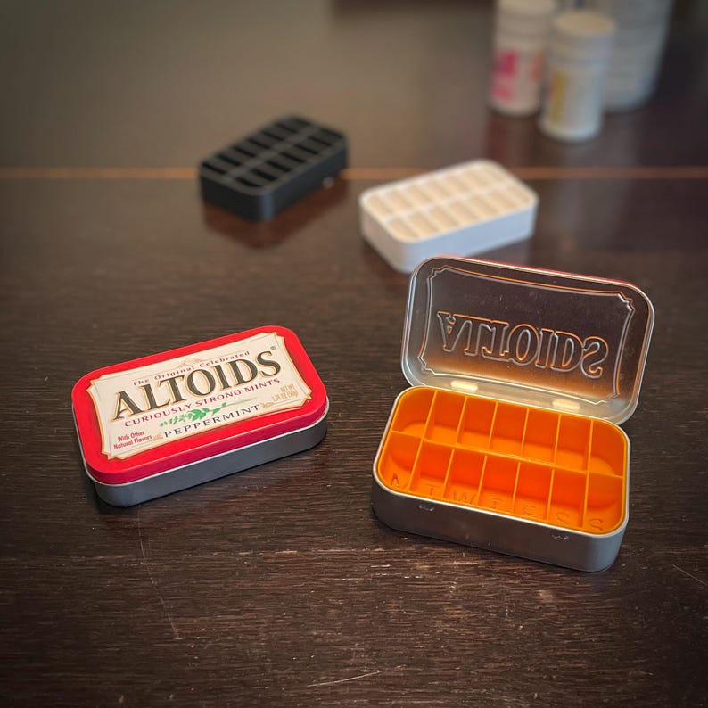 Puede incluir: Una lata de Altoids roja y plateada con el texto "The Original Celebrated Altoids Curiously Strong Mints Peppermint" en la tapa. La lata est&aacute; abierta y revela un organizador de p&iacute;ldoras naranja con 12 compartimentos.