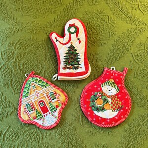 May include: Three Christmas-themed kitchen textiles: an oven mitt and two pot holders. The oven mitt is white with a red trim and features a Christmas tree design. The pot holders have a gingerbread house and snowman design.