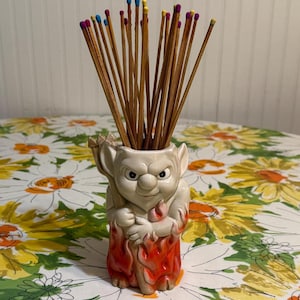 May include: A white ceramic matchstick holder shaped like a devil figure with red flames. The devil is holding a pitchfork and has a mischievous expression. The holder is filled with wooden matchsticks with colorful tips.