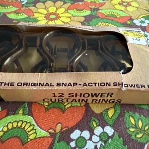 May include: A package of 12 brown snap-action shower curtain rings. The cardboard box has the text "THE ORIGINAL SNAP-ACTION SHOWER" and "12 SHOWER CURTAIN RINGS". The background is a floral pattern with orange, yellow, and green flowers.