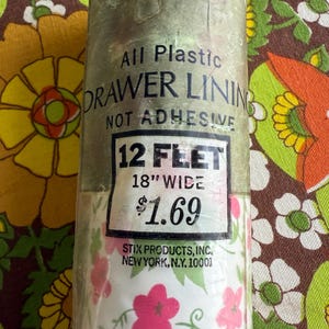 May include: A roll of clear plastic drawer lining with a floral pattern of pink flowers and green leaves on a white background. The label reads "All Plastic Drawer Lining, Not Adhesive, 12 Feet, 18" Wide."