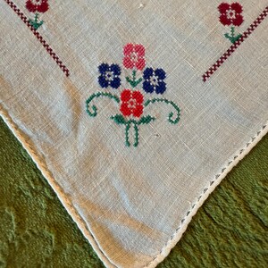 May include: Close-up of a vintage, off-white linen handkerchief with embroidered floral designs. The embroidery features red, pink, and blue flowers with green stems and accents. The edges are finished with a delicate, white, stitched border.