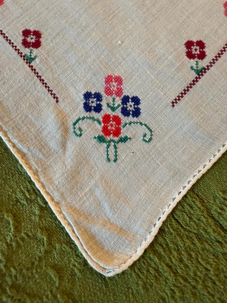 May include: Close-up of a vintage, off-white linen handkerchief with embroidered floral designs. The embroidery features red, pink, and blue flowers with green stems and accents. The edges are finished with a delicate, white, stitched border.