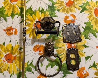 May include: A collection of antique brass bathroom fixtures, including a towel bar, toilet paper holder, towel ring, and light switch covers. The fixtures have ornate designs and are arranged on a floral patterned background.