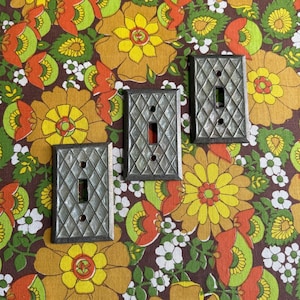 May include: Three vintage light switch covers with a diamond pattern. The covers are silver-toned with a rectangular opening for the switch and two screw holes. The switches have a red toggle. The background is a floral pattern in shades of brown, yellow, orange, and green.