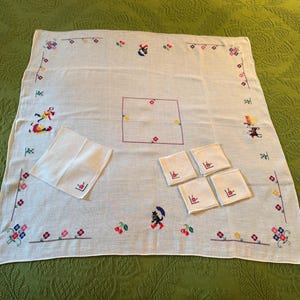 May include: White embroidered tablecloth with matching napkins and coasters. The tablecloth features colorful floral and whimsical figure embroidery around the edges and center. The set includes a large square tablecloth, a napkin, and four coasters.