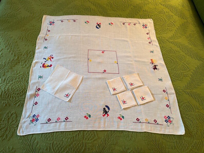 May include: White embroidered tablecloth with matching napkins and coasters. The tablecloth features colorful floral and whimsical figure embroidery around the edges and center. The set includes a large square tablecloth, a napkin, and four coasters.