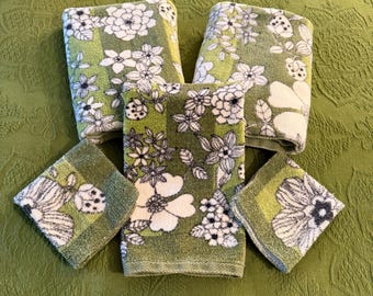 May include: A set of six green and white floral patterned towels and washcloths. The towels and washcloths have a repeating pattern of large white flowers with black and gray accents. The background is a textured green fabric.