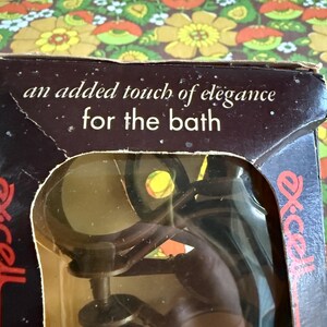 May include: A vintage bathroom accessory in its original packaging. The box is brown with the text "an added touch of elegance for the bath." The accessory is visible through a clear plastic window.