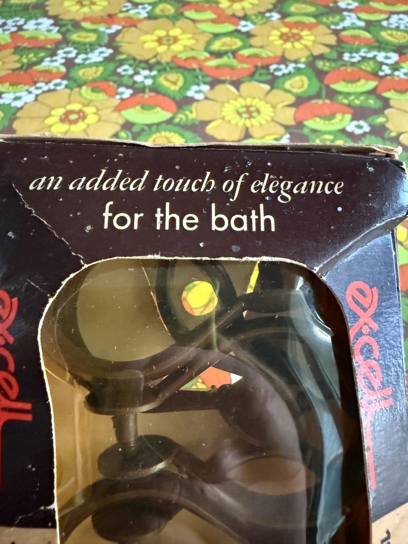 May include: A vintage bathroom accessory in its original packaging. The box is brown with the text "an added touch of elegance for the bath." The accessory is visible through a clear plastic window.