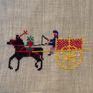 May include: Cross-stitch embroidery of a horse-drawn cart with a driver. The horse is black with red accents, pulling a yellow and red cart. The driver wears blue and purple. The background is a light beige fabric.