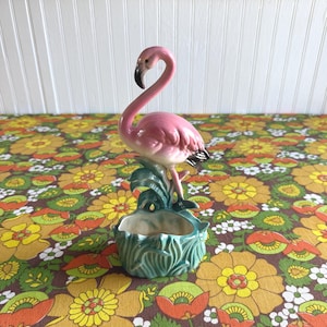 May include: A ceramic flamingo figurine planter featuring a pink flamingo with a black-tipped beak standing atop a glossy teal green base molded to resemble marshy foliage and reeds, with a hollow opening at the front for small plants.