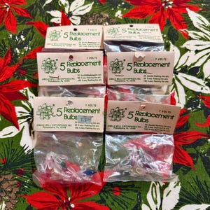May include: Several packages of replacement Christmas light bulbs are displayed on a festive floral patterned fabric. Each package contains five bulbs and is labeled with the text "Jingle Bell" and "5 Replacement Bulbs". The bulbs are clear with red, blue, and green accents.