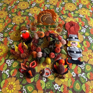 May include: A collection of Thanksgiving-themed items including a turkey napkin holder, two small glasses, a fruit and leaf wreath, three plush turkey ornaments, a small turkey figure, and a felt wall hanging depicting a pilgrim figure.