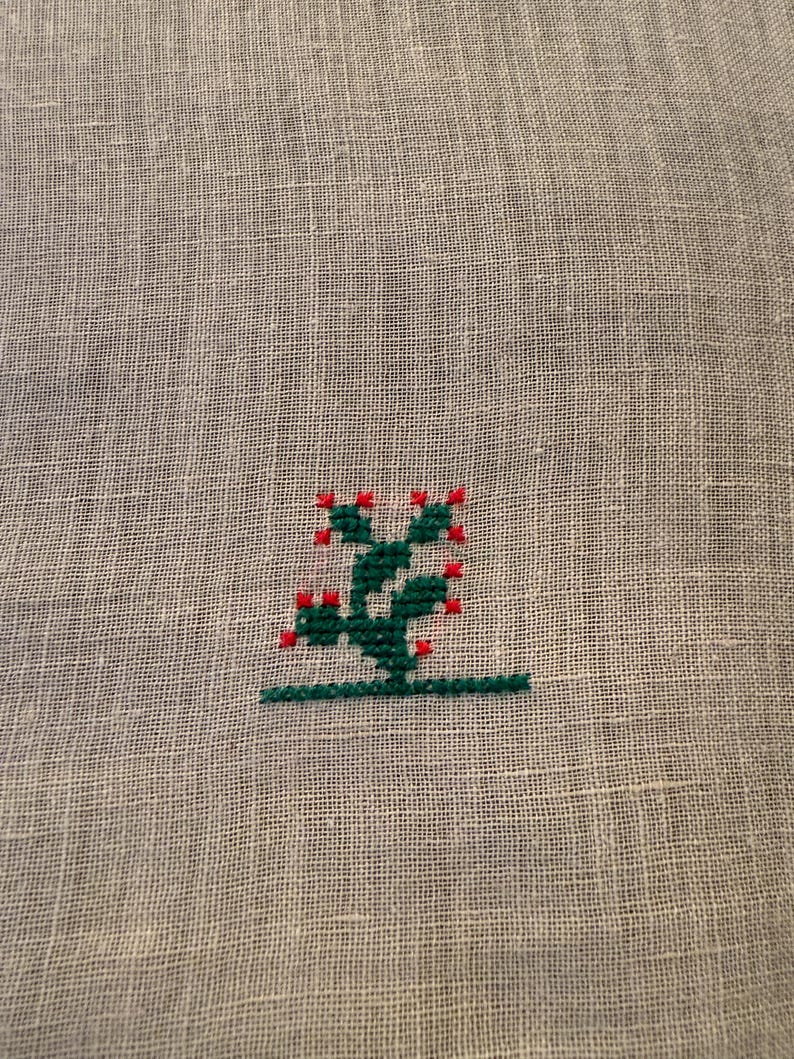 May include: A close-up of a gray linen fabric with a small, embroidered cactus design. The cactus is green with red accents, and the embroidery is done in a cross-stitch style. The design is centered on the fabric.