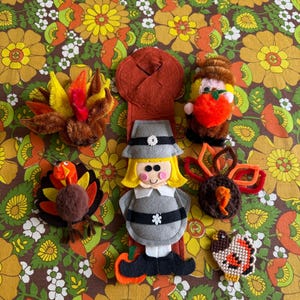 May include: A collection of Thanksgiving-themed felt and yarn ornaments, including turkeys and a pilgrim figure. The ornaments are in various colors, including brown, orange, yellow, and gray, and are arranged on a floral patterned background.