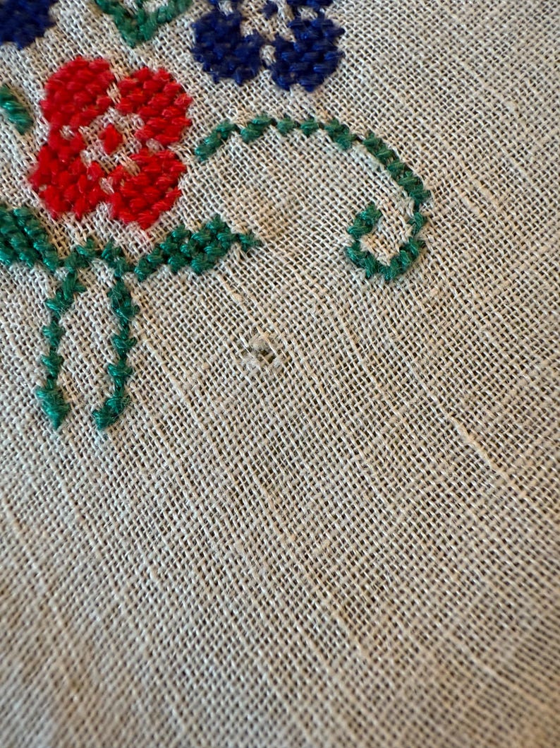 May include: Close-up of a beige linen fabric with embroidered floral details. The embroidery features red, blue, and green thread, forming flowers and leaves. The fabric has a visible woven texture.