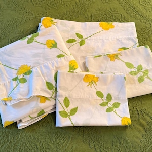 May include: White bed sheets and pillowcases with a yellow rose floral pattern. The roses have green stems and leaves. The bedding is arranged on a green textured surface.