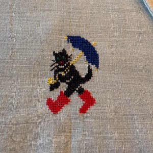 May include: A cross-stitched design of a black cat wearing red boots and holding a blue umbrella with yellow accents. The cat has green eyes and a yellow collar. The design is on a light beige linen fabric.